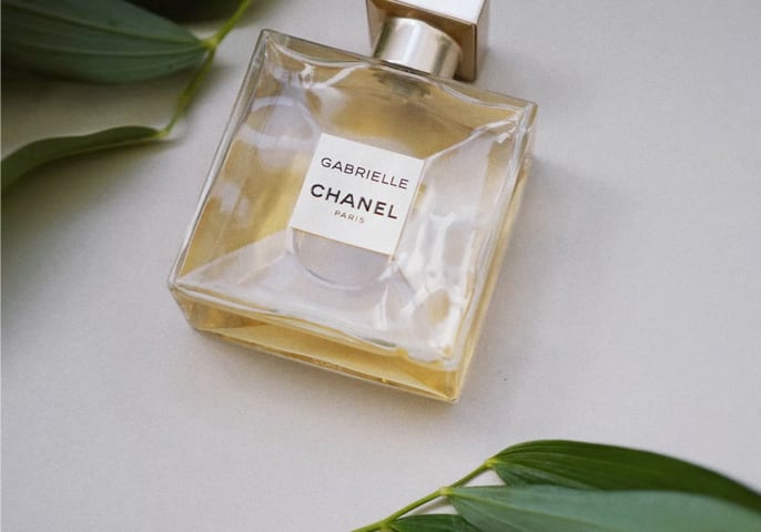 Gabrielle Essence Eau De Parfum Bottle Lying on a flat
        surface with green leaves below and above the bottle